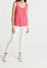 Lyserød ærmeløs tanktop med rund halsudskæring og lille brystlogo, parret med tætsiddende hvide jeans og høje sneakers.