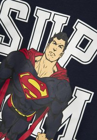 Marineblauw t-shirt met een afbeelding van een gespierde superheld in een blauw en rood kostuum met een gele embleem, naast grote witte letters die "SUP" zeggen.