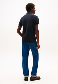 Tamsiai mėlynas polo marškinėlis iš medvilnės su klasikiniu apykakle, derinamas su šviesiai mėlynais tiesiais džinsais ir rudais batais.