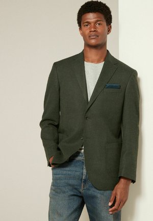 Dark green blazer with a textured fabric, notch lapels, two buttons, and a chest pocket containing a blue handkerchief. Worn over a grey shirt.