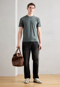 Grå bomulls t-skjorte med korte ermer og hvit tekst, kombinert med mørke jeans og grønstripete sneakers, holder en brun lærveske.