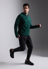 Pullover de fleece verde com colarinho com fecho e punhos, combinado com calças de desporto pretas com riscas laterais e sapatilhas pretas.