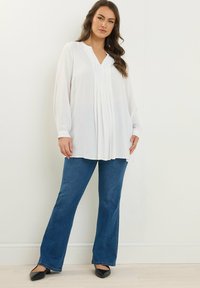 Camicia bianca a maniche lunghe con scollatura a V e pieghe verticali, abbinata a jeans a campana blu e scarpe nere, su uno sfondo semplice.