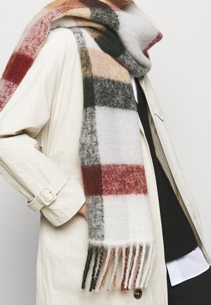 Personne portant un trench-coat crème et une écharpe en laine à carreaux multicolores avec franges, par-dessus un pull noir et une chemise blanche.