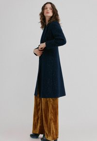 Femme portant un long manteau bleu marine à motifs, par-dessus un pantalon large en velours moutarde, debout de profil avec les mains près de la taille.