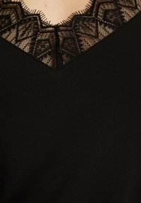 Maglietta nera con scollo a V adornato da dettagli in pizzo nero intricati e bordi ondulati, che evidenziano il contrasto di texture.