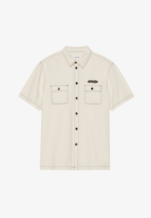 Camisa de algodão leve de mangas curtas na cor bege, com botões pretos, dois bolsos no peito e costuras contrastantes em preto. Logótipo no bolso esquerdo.