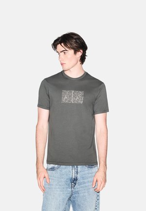 Giovane uomo con maglietta grigia con grafica e jeans azzurri chiari che guarda di lato, in piedi contro uno sfondo chiaro e uniforme.