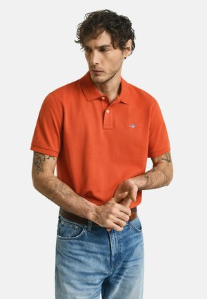 Kortærmet orange polo-shirt med krave, som har et lille logo på brystet. Sammen med lyseblå jeans og et brunt bælte.