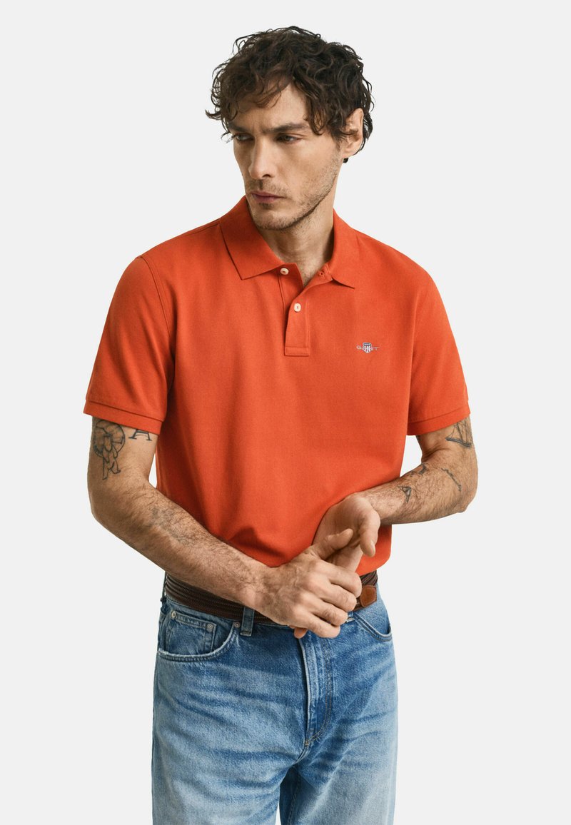 Kortærmet orange polo-shirt med krave, som har et lille logo på brystet. Sammen med lyseblå jeans og et brunt bælte.