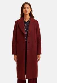 Femme portant un long manteau bordeaux avec de grands revers sur une chemise à motif sombre, se tenant devant un fond blanc uni.