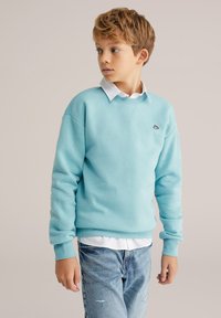 Svetlomodrá mikina s okrúhlym výstrihom a rebrovanými manžetami, vrstvená cez bielu košeľu s golierom. Nosí sa s vyblednutými modrými džínsami.