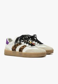 Sneaker bianche con strisce leopardate, lacci trasparenti neri, linguette color viola sul tallone e decorazioni a ciondolo su suole marrone chiaro.