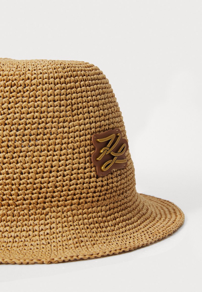 Hellbrauner, geflochtener Strohhut in Form eines glatten, abgerundeten Designs mit einem braunen Lederpatch, das ein geprägtes goldfarbenes Logo zeigt.