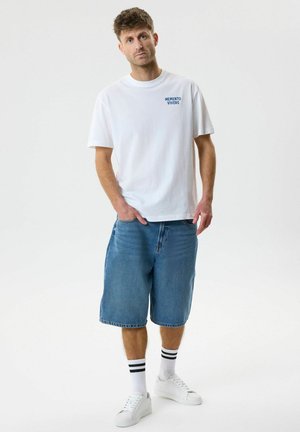 Ung mann står med hendene i lommene, iført hvit "Memento Vivere" T-skjorte, lange denimshorts, hvite sokker med svarte striper og hvite joggesko.