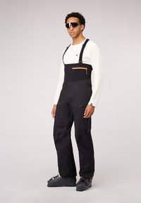 Schwarze Schnee-Latzhose mit verstellbaren Trägern, orangefarbigem Reißverschlussfach und seitlichen Reißverschlüssen. Getragen über einem weißen Langarmshirt und kombiniert mit grauen Skischuhen.