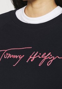 Felpa nera con collo rotondo. Presenta ricamata in rosso la firma "Tommy Hilfiger" in un font corsivo. Tessuto casual e testurizzato.