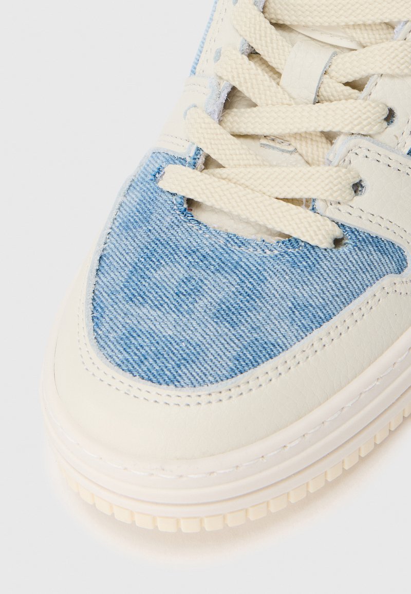 Sneaker di pelle bianca con pannelli in denim azzurro chiaro, superficie testurizzata e lacci beige. Presenta una punta arrotondata e una suola ammortizzata.
