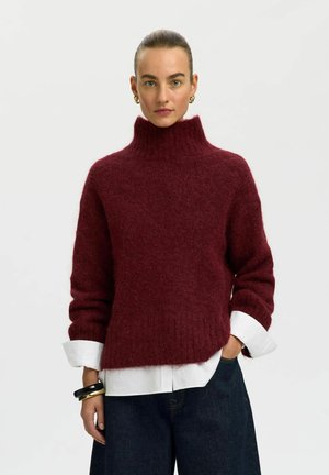 Mujer con un suéter de cuello alto burdeos sobre una camisa blanca, jeans azul oscuro, pendientes de aro dorados y pulseras en la muñeca izquierda