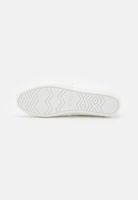 Hvit sneakersåle med teksturert gummi for grep. Har et sikksakkmønster og preget "TOMS"-merke nær hælen. Glatt finish.