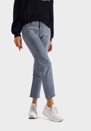 Persona con jeans azzurri chiari, felpa blu navy e sneaker bianche senza lacci in piedi con una gamba leggermente incrociata su uno sfondo semplice.