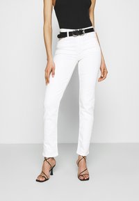 Los jeans blancos de pierna recta presentan una cintura de media altura, combinados con un cinturón de cuero sintético negro con una hebilla circular y tacones negros con tiras.