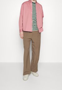Rosa Bluse über einem grün-weißen gestreiften T-Shirt, kombiniert mit braunen weiten Hosen und weißen Sneakers.