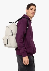 Svetle šedý batoh s několika zipy a přední kapsou, nošený přes tmavě fialovou mikinu s kapucí. Obsahuje nastavitelné popruhy a m�á strukturovaný tvar.