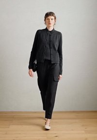 Cárdigan de punto negro con botones, llevado sobre una camisa oscura y pantalones cargo. El modelo usa zapatos claros y lleva un bolso pequeño negro. Suelo de madera.