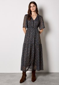 Długa, zwiewna granatowa sukienka z wielokolorowym wzorem fal, krótkimi rękawami i wiązaniem w pasie; zestawiona z brązowymi botkami na obcasie.