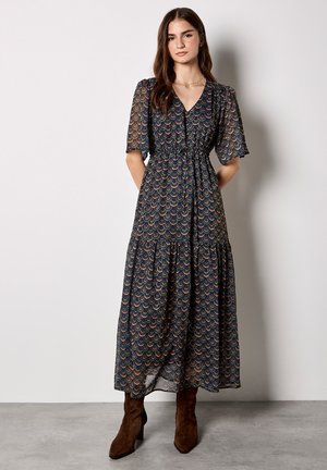 Lang, vloeiende marineblauwe jurk met een veelkleurig golfpatroon, korte mouwen en een strik taille; gecombineerd met bruine haklaarzen.