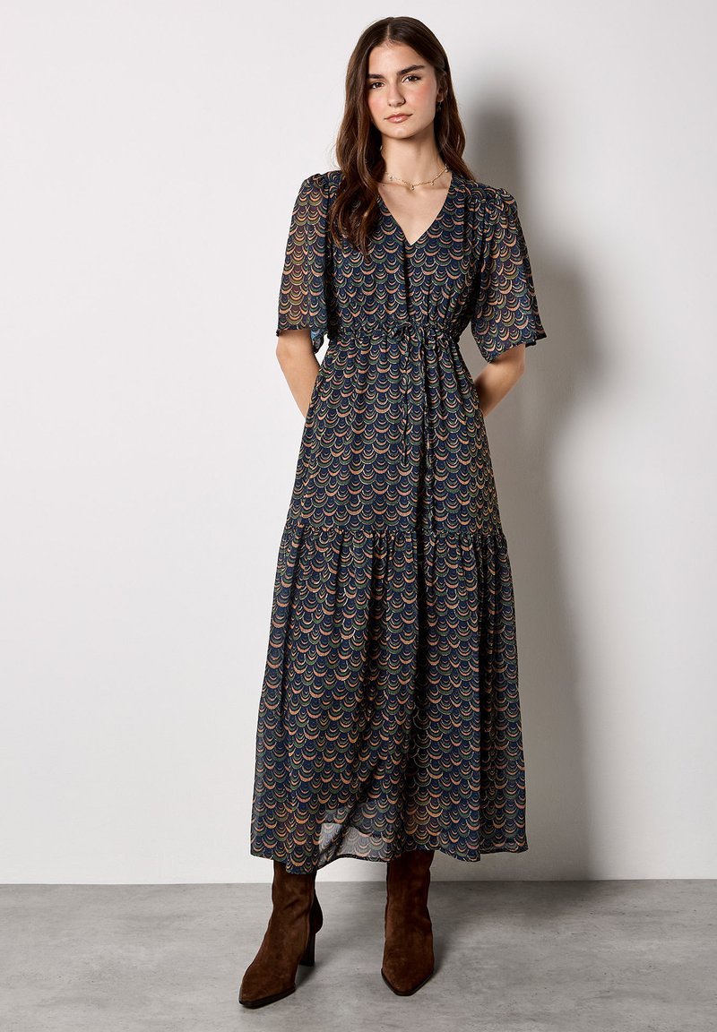 Długa, zwiewna granatowa sukienka z wielokolorowym wzorem fal, krótkimi rękawami i wiązaniem w pasie; zestawiona z brązowymi botkami na obcasie.