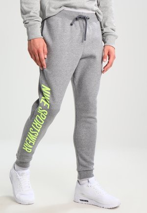 Mann trägt graue Jogginghose mit neongelbem Schriftzug „NIKE SPORTSWEAR“ am linken Bein und weiße Nike-Sneaker, steht vor hellem Hintergrund.