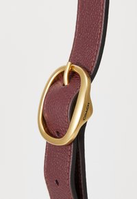 Burgundy leather belt with a textured finish, featuring a gold-tone metal buckle and a smooth black lining. Brand name embossed on hardware.