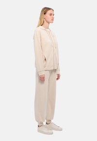 Felpa con cappuccio beige e pantaloni della tuta realizzati in tessuto morbido, dotati di chiusura con zip, tasche laterali e polsini affusolati, abbinati a sneakers color crema.