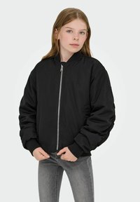 Črna bomber jakna z zadrgo, rebrastimi manšetami in sproščenim krojem. Izdelana iz gladkega blaga. Kombinirana s sivimi oprijetimi kavbojkami.