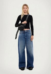 Woman wearing black long-sleeve top with eyelet details, wide-leg blue jeans, black shoes, and a black crossbody bag with a grommet strap.