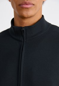 Chaqueta negra con cierre y cuello alto, hecha de un tejido suave, que presenta un cuello alto y un detalle de costura mínima en la parte delantera.