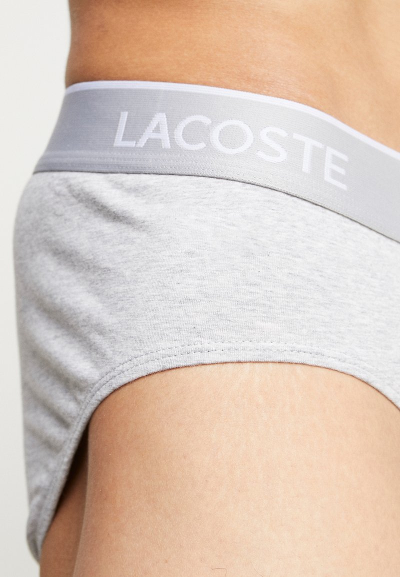 Bañador gris de algodón con una cinturilla gris claro que lleva "LACOSTE" en letras blancas. El material tiene una textura suave y un diseño casual.