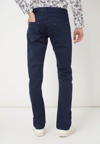 Hombre de pie con una mano en el bolsillo trasero, vestido con pantalones azul oscuro, zapatillas blancas y una camisa blanca con patrones de hojas negras.