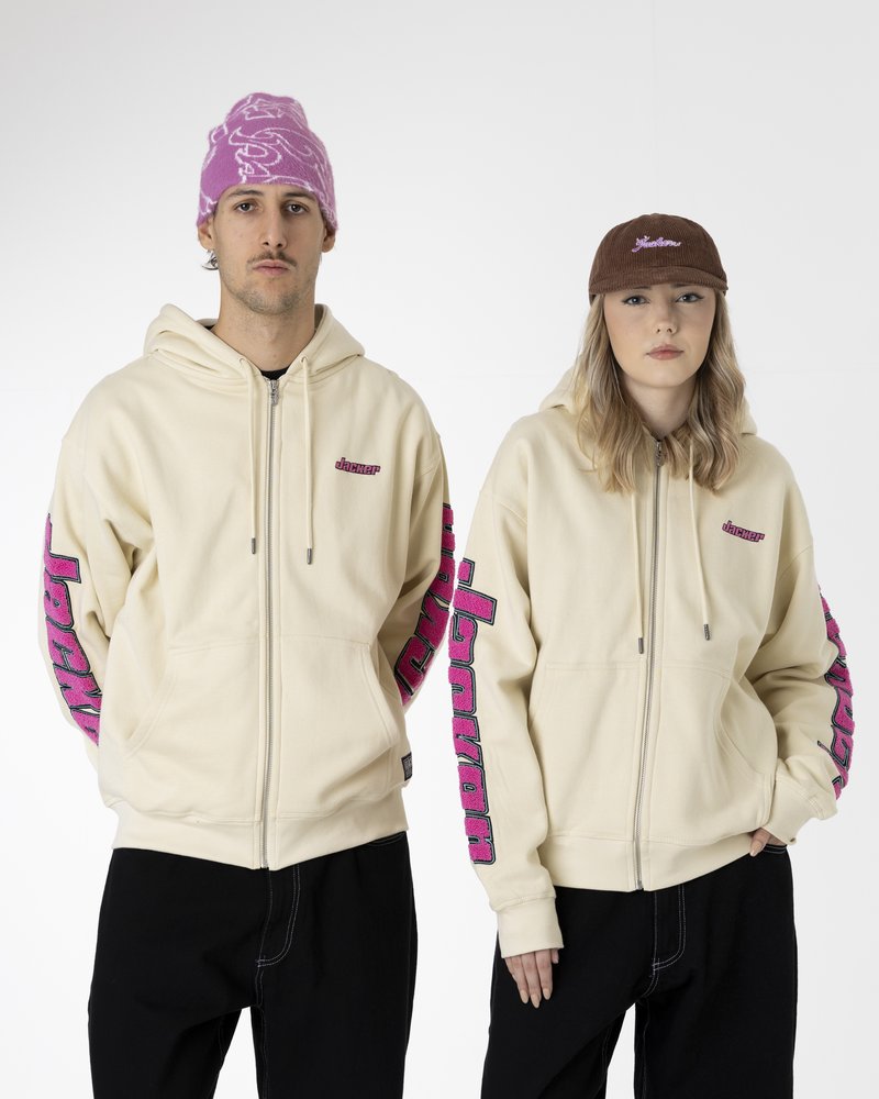 Krém színű zipzáros kapucnis pulóver rózsaszín grafikai szöveggel az ujjak mentén, sötét nadrággal párosítva. Rózsaszín sapka és barna kalap is látható.
