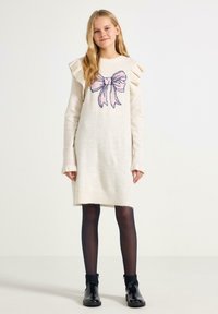 Crèmefarvet strikkjole med rynkede skuldre og et pink bue-grafik, parret med sorte strømpebukser og skinnende sorte ankelstøvler.