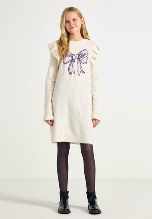 Krämfärgad stickad klänning med volangärmar och en rosa rosettgrafik, kombinerad med svarta strumpbyxor och blanka svarta ankelskot.