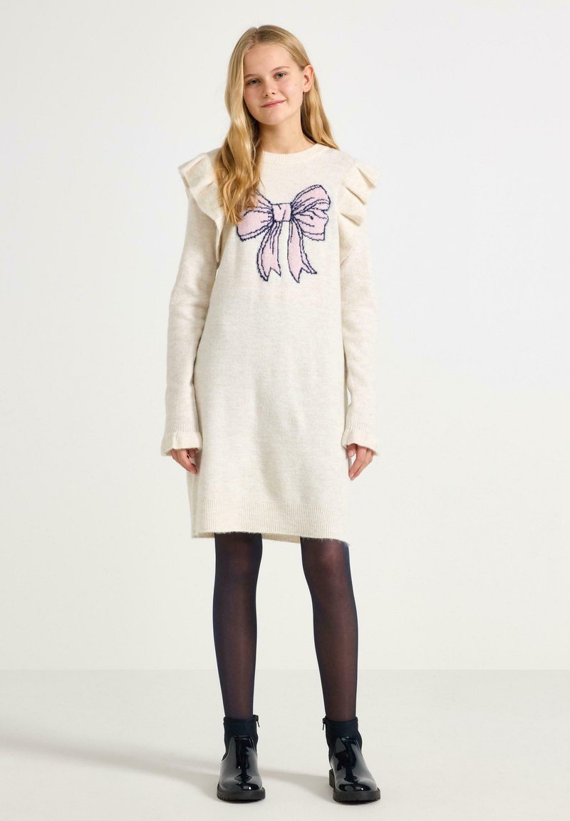 Crèmefarvet strikkjole med rynkede skuldre og et pink bue-grafik, parret med sorte strømpebukser og skinnende sorte ankelstøvler.