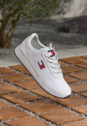Weißer Sneaker mit rotem, weißem und blauem Tommy Jeans Logo, der über hölzernen quadratischen Fliesen im Freien mit einem Baum und Kies im Hintergrund schwebt.