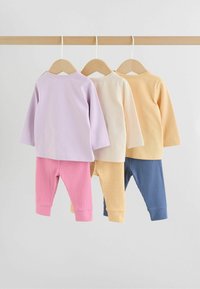 Tres conjuntos de ropa de bebé cuelgan en perchas de madera, cada uno con una camiseta de manga larga y pantalones de canalé en púrpura pastel, crema y amarillo.