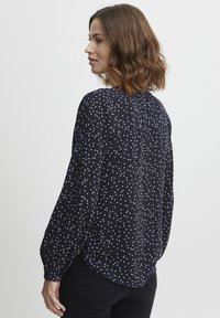 Marineblaue Bluse mit weißen Punkten, langen Ärmeln mit elastischen Bündchen, abgerundetem Saum und aus leichtem Stoff gefertigt. Model seitlich positioniert.