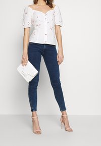 Vit blus med puffärmar och röd blommig broderi, knäppning framtill; matchad med mörkblå skinny jeans och blekrosa öppna klackar. Håller en vit kuvertväska.