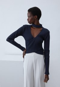 Top de canalé navy con diseño envolvente y cuello alto; combinado con pantalones anchos blancos con franjas laterales negras.