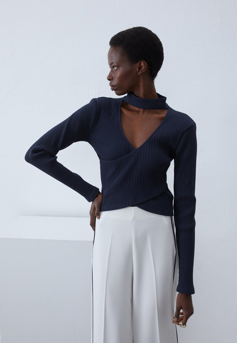 Top de canalé navy con diseño envolvente y cuello alto; combinado con pantalones anchos blancos con franjas laterales negras.